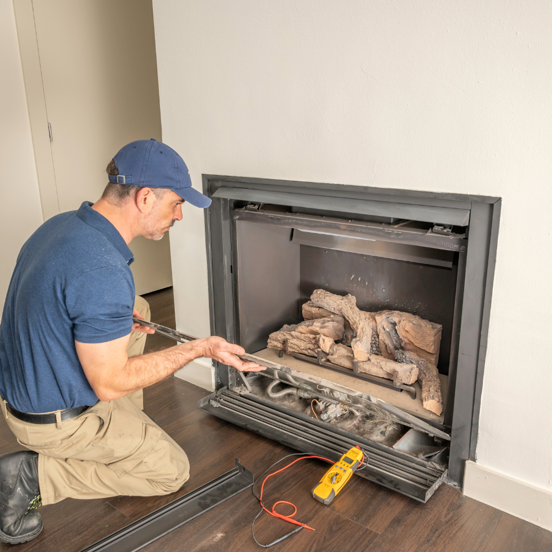 Man in blue shirt and cap kneeling, inspecting a gas fireplace with tools.