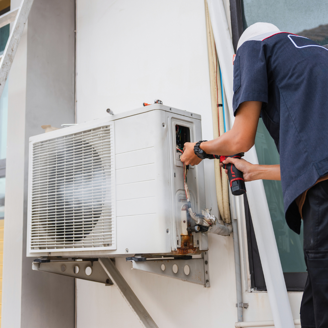 HVAC technician using a drill to repair an outdoor air conditioning unit mounted on a wall.