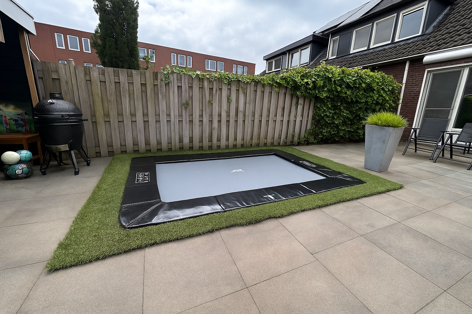 Een rechthoekige, ingegraven trampoline in een achtertuin, omgeven door kunstgras en bestrating.
