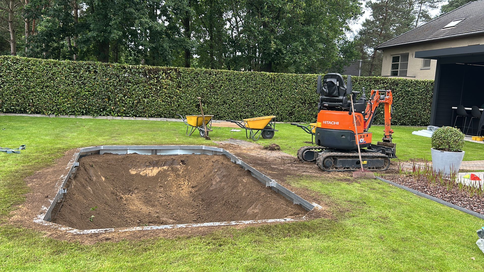 Een minigraafmachine graaft een rechthoekig gebied uit in een grasrijke achtertuin, omgeven door struiken en een huis.