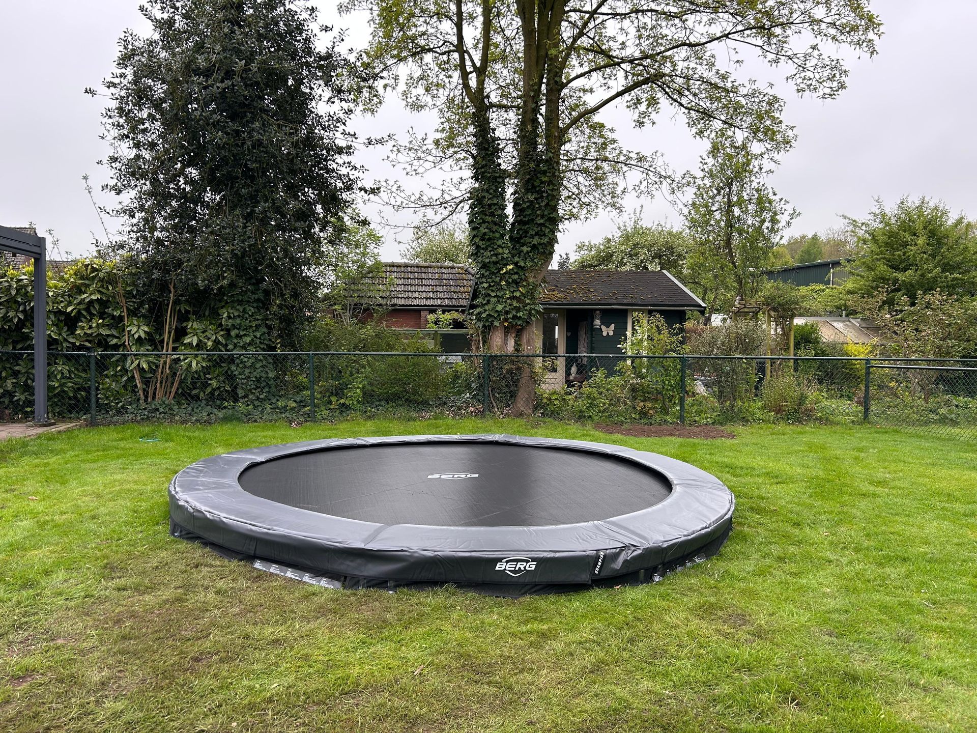 Ronde zwarte trampoline in een grasveld, vlakbij bomen en een hek.