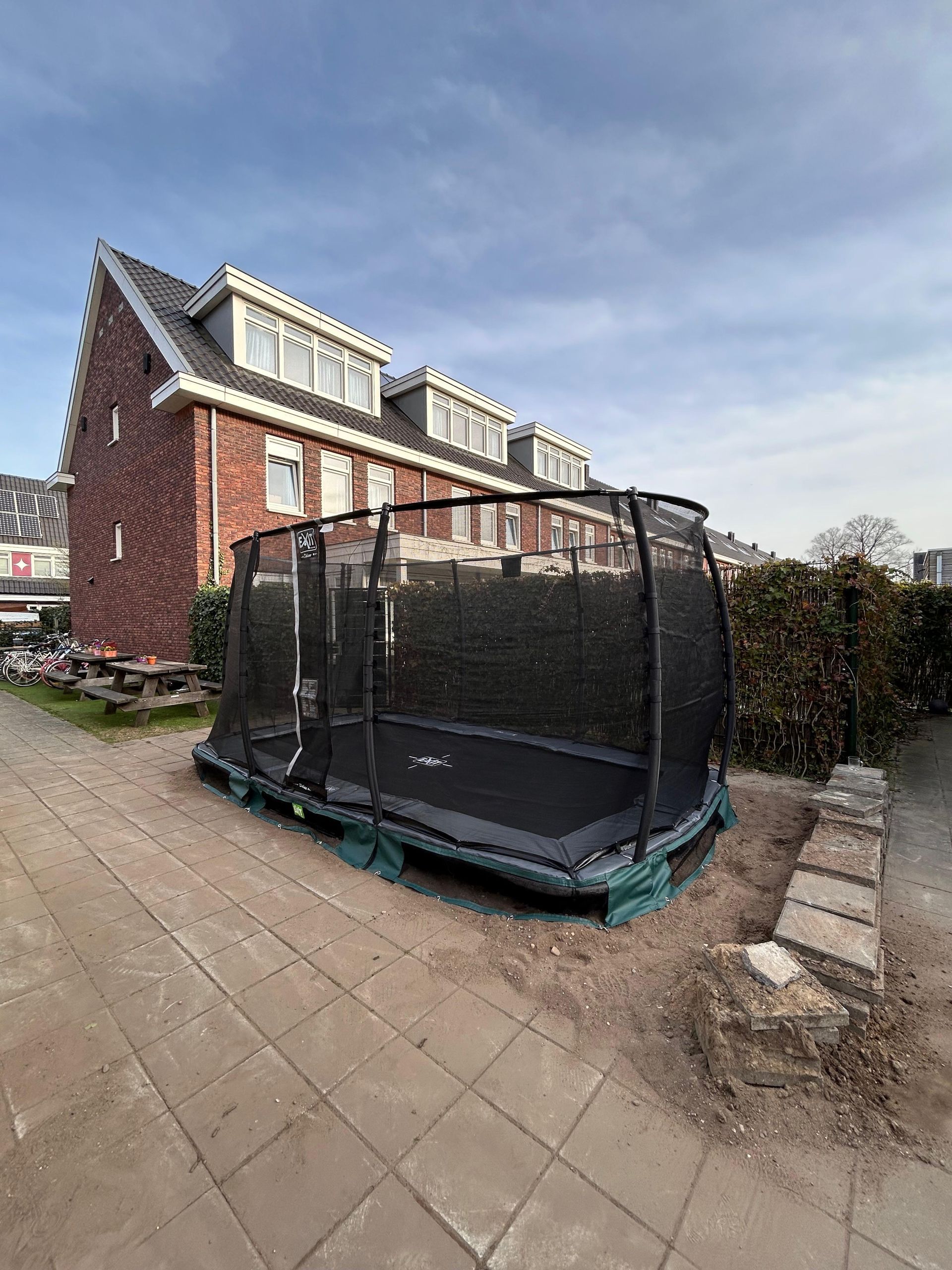 Een trampoline in een achtertuin, vlakbij een bakstenen huis, onder een bewolkte hemel.