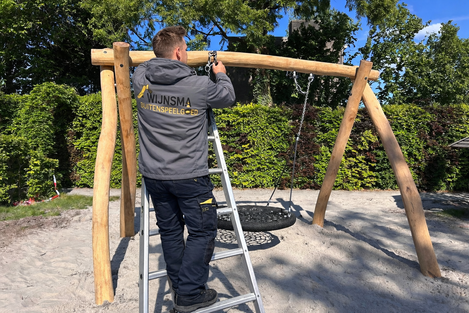 Een man staat op een ladder om een ​​bandenschommel te bevestigen aan een houten schommelset in een zanderige speelruimte.