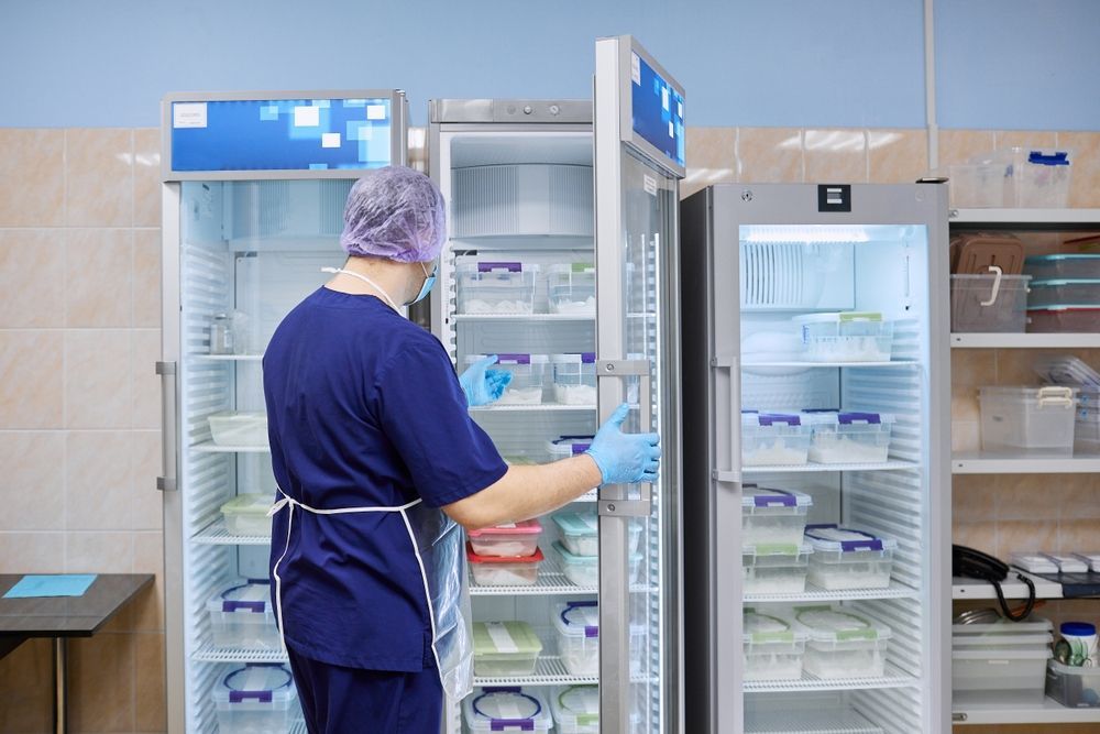 Person in scrubs opening refrigerator in a lab, filled with containers and supplies.
