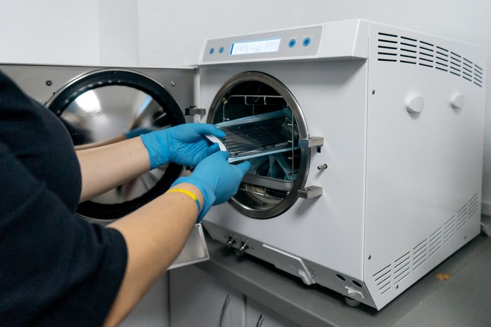Person in blue gloves placing a tray of instruments into a sterilizer. White machine in a clinic setting.