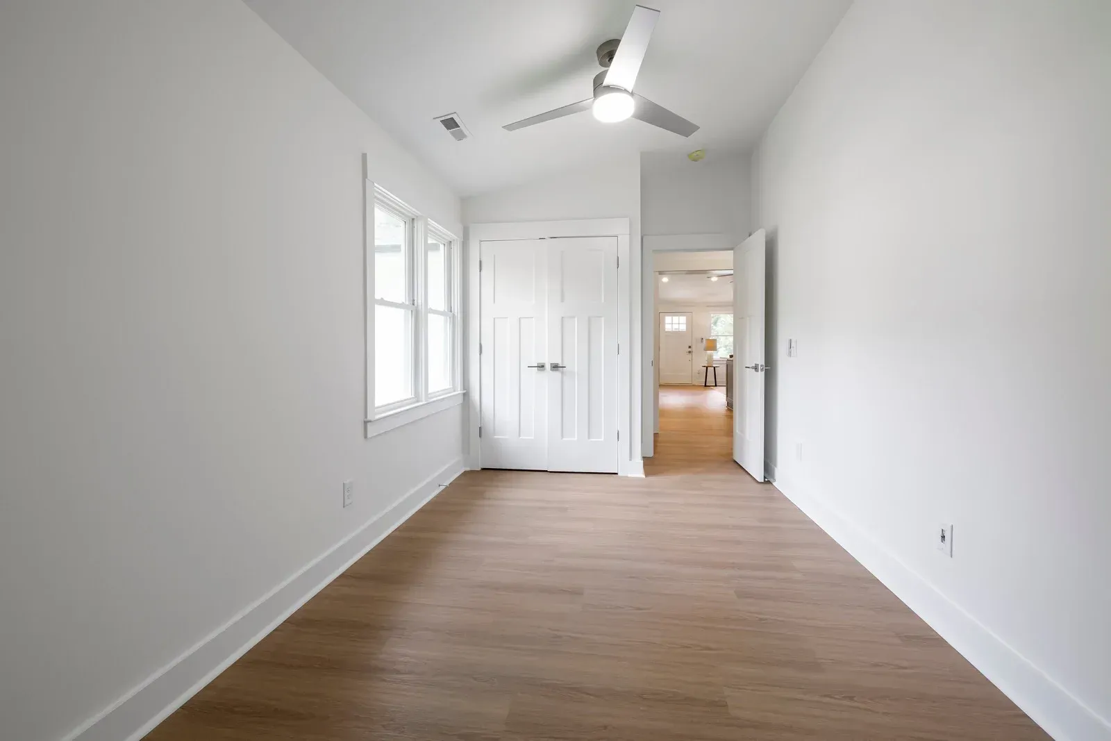 Empty bedroom with white walls, closet, window, and wood-look floor; ceiling fan.