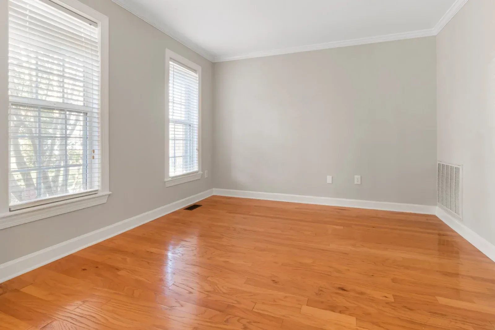 Empty room with hardwood floors, two windows with blinds, and light gray walls.