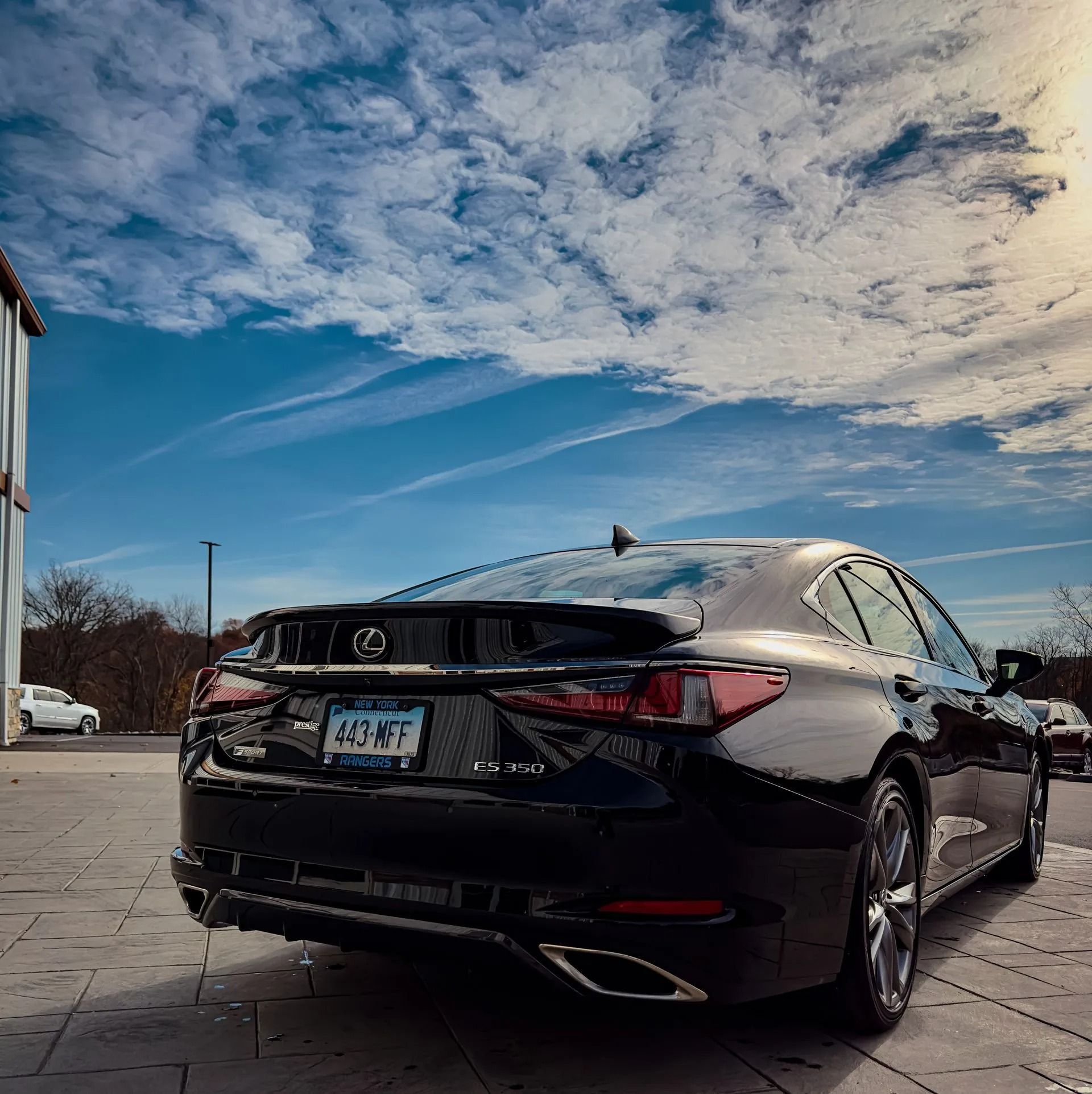 A black Lexus sedan parked on a paved lot under a blue sky with scattered clouds.