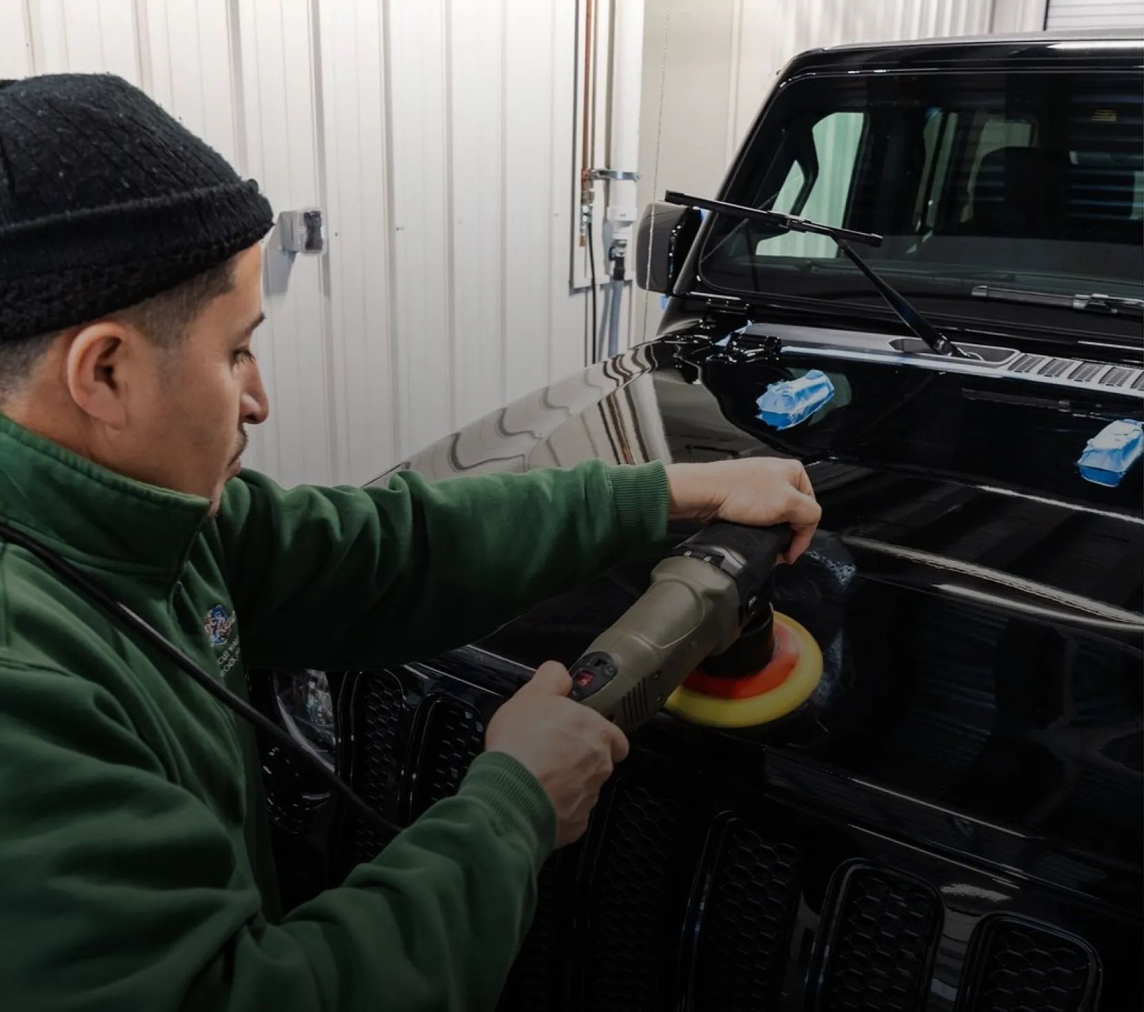 A person in a green fleece and beanie uses a power polisher on the hood of a black vehicle in a garage.