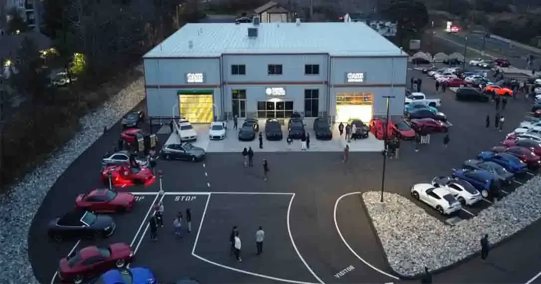An aerial view of a car meet at a commercial building with many parked vehicles and people standing in the lot at dusk.