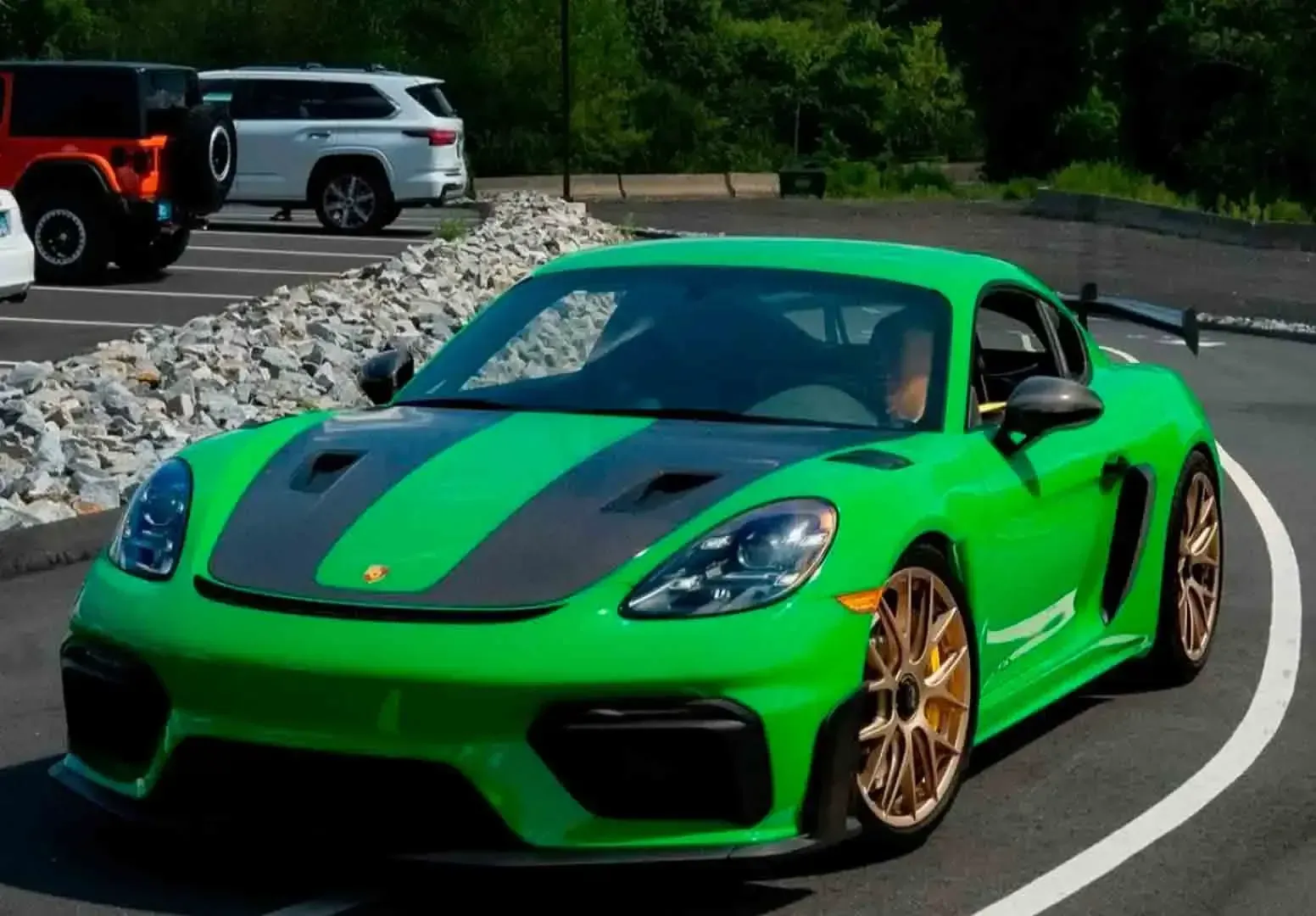 A bright green Porsche 718 Cayman GT4 RS with a black hood and gold wheels parked on a paved road next to other vehicles.
