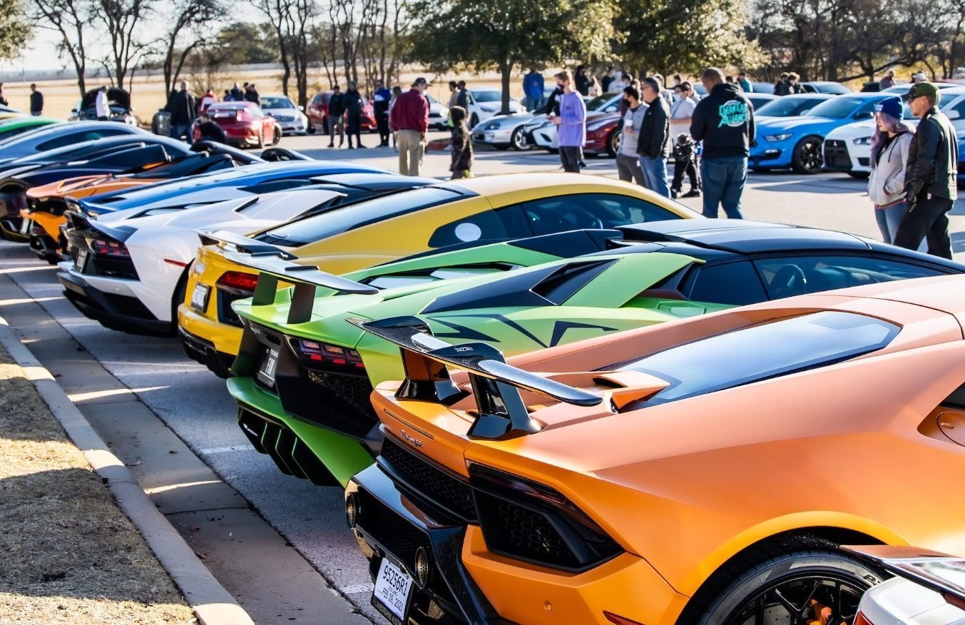 A line of colorful Lamborghini sports cars parked outdoors at a sunny gathering with people walking nearby.