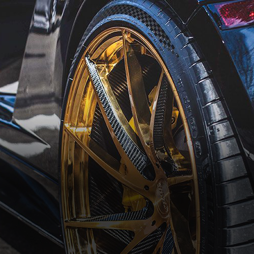 A gold-rimmed car wheel with carbon fiber spokes and a dark tire, viewed from a close-up angle.