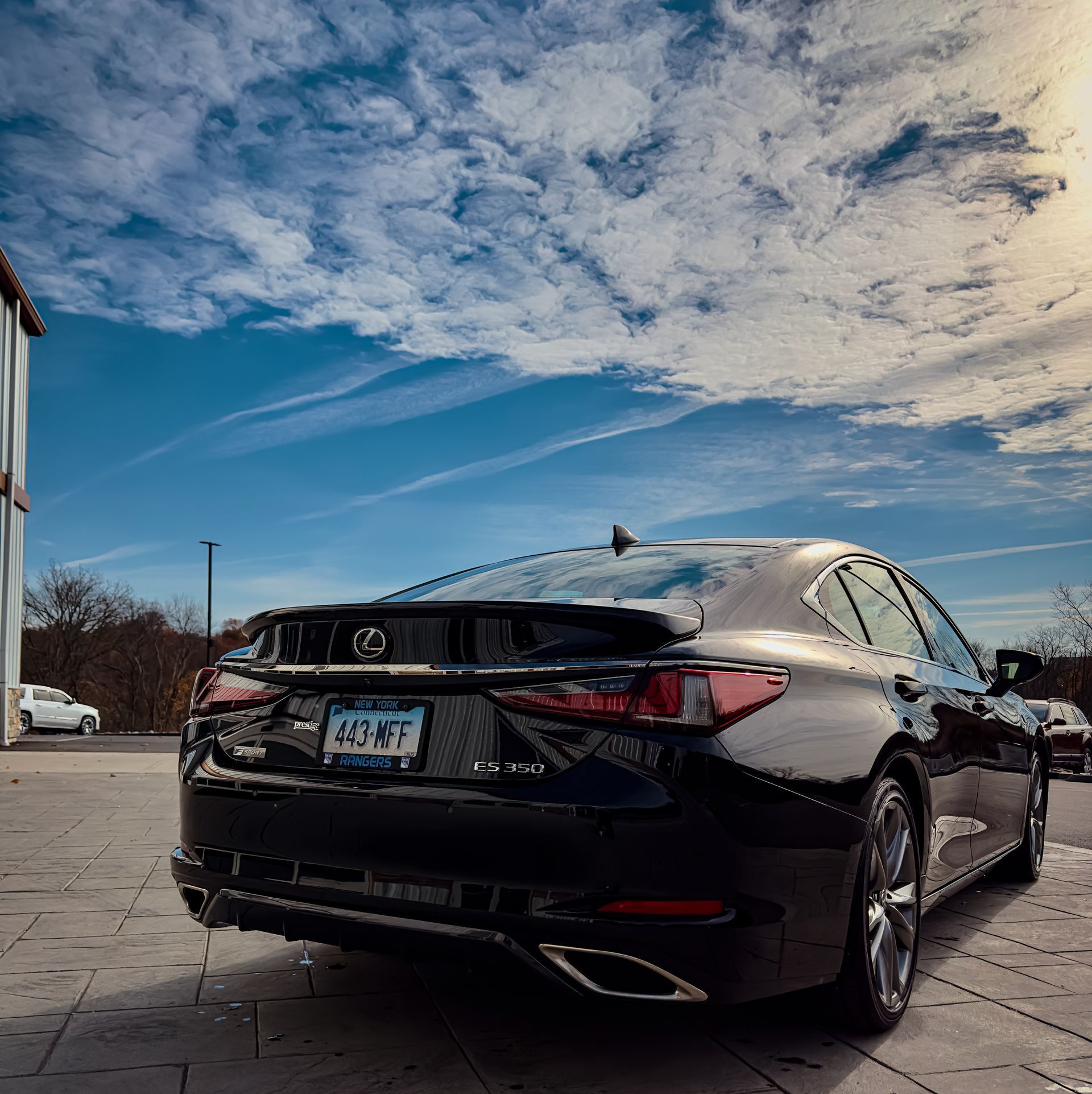 A black Lexus sedan parked on a paved lot under a bright blue sky with wispy clouds.