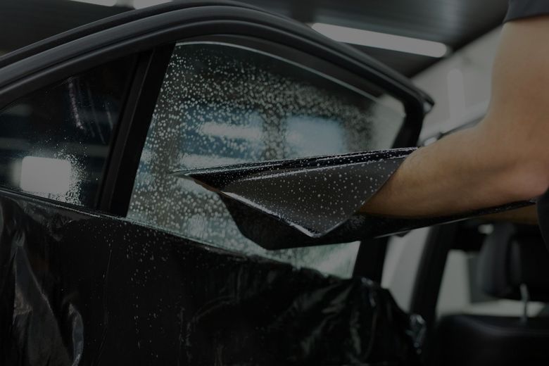 A person carefully applies a dark window tint film to a car window that is covered in soapy water.