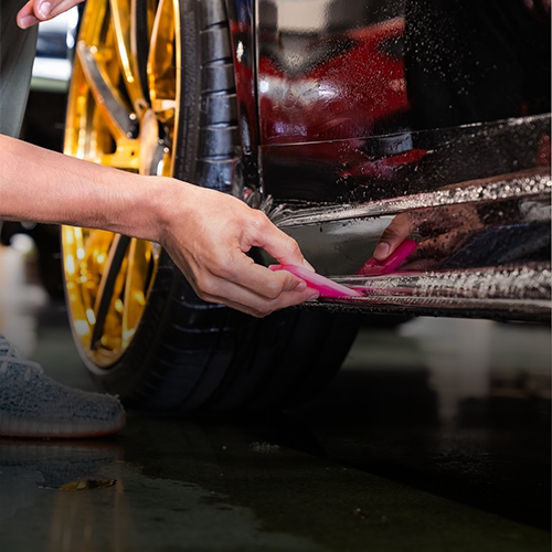 A technician uses a pink squeegee to smooth a protective film onto the side panel of a car with gold-rimmed tires.