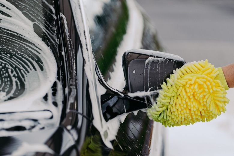 A yellow microfiber mitt washes the side mirror of a black car covered in white soap suds.