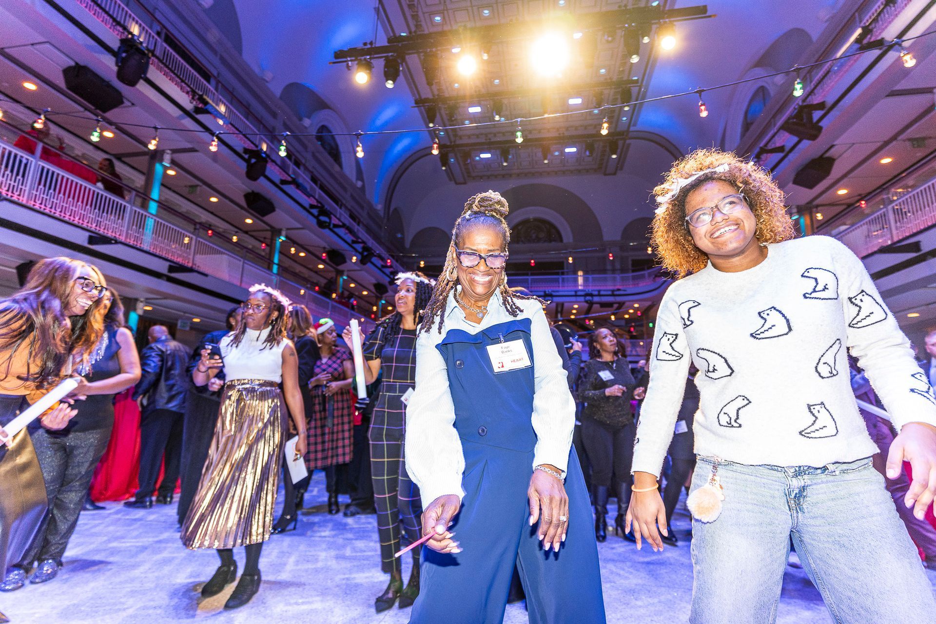 HOS Holiday Celebration - People dancing in a large hall with stage lighting. Some wear party attire, holding glow sticks, smiling.