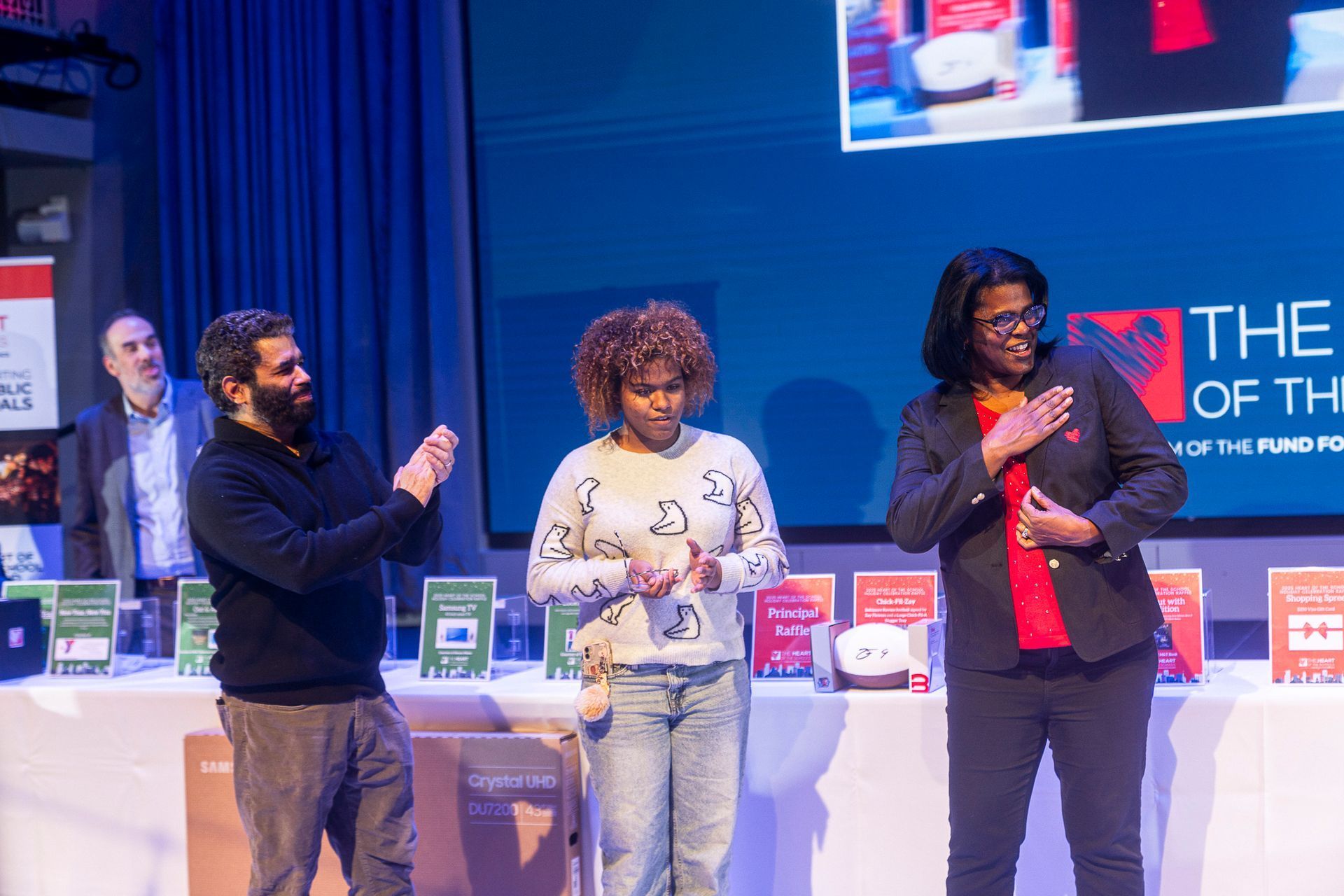 HOS Holiday Celebration - Three people on stage, awards event. One applauds, two look down, illuminated by blue light.