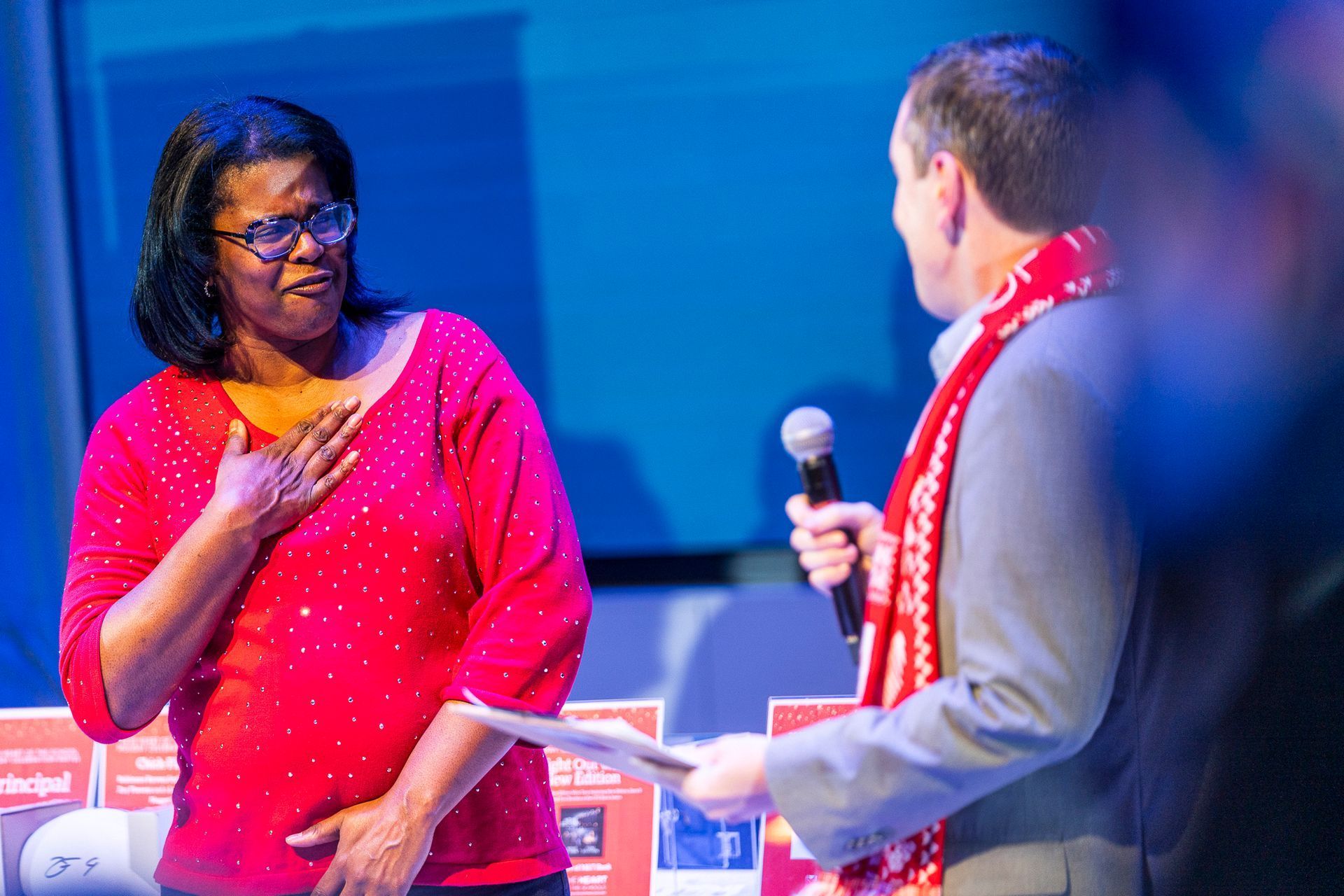 HOS Holiday Celebration - Woman in red top and glasses reacts as man with microphone speaks; stage setting.