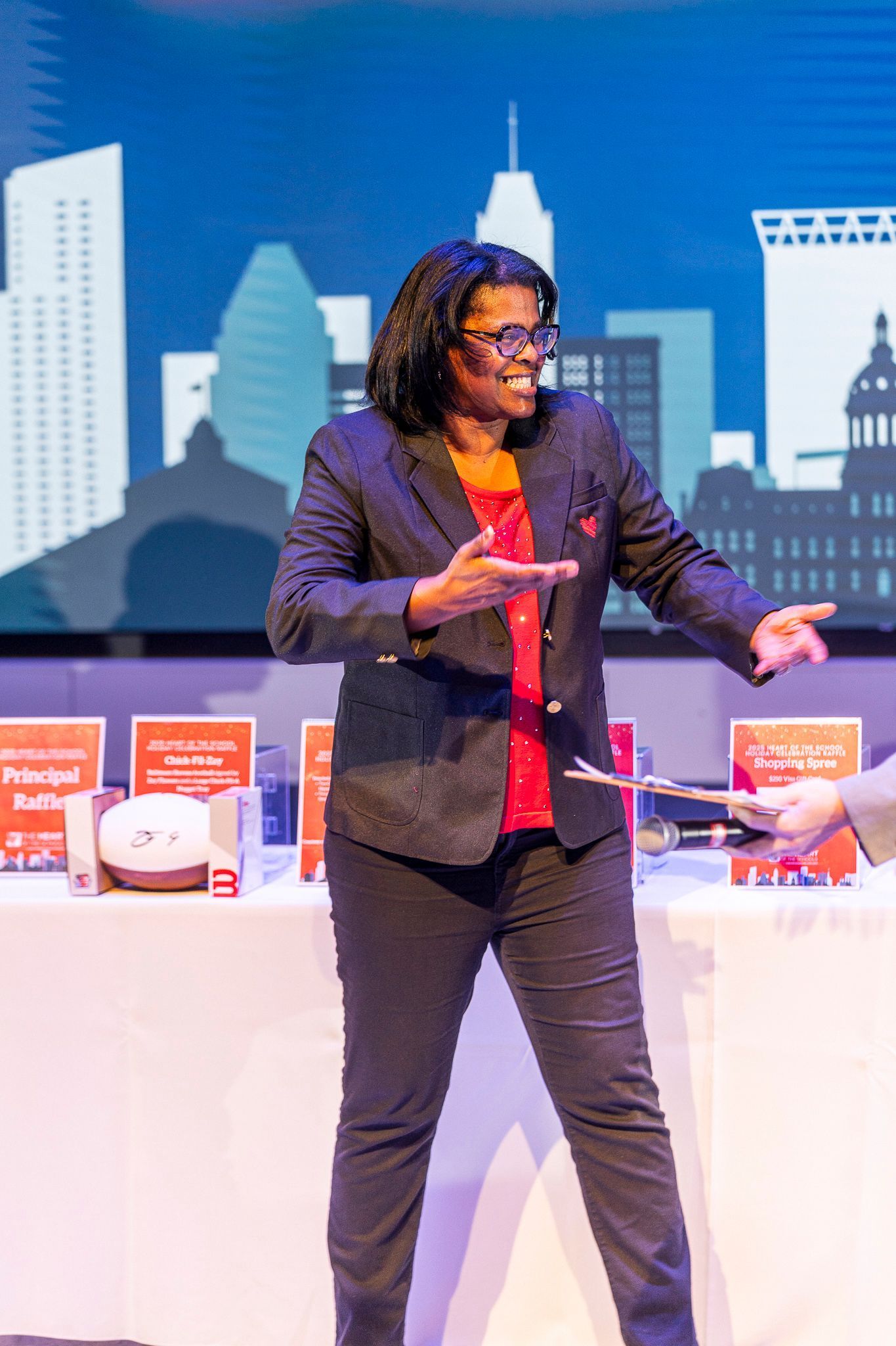 Woman pointing, smiles. On stage with cityscape backdrop, wearing a blazer over a red shirt, black pants.