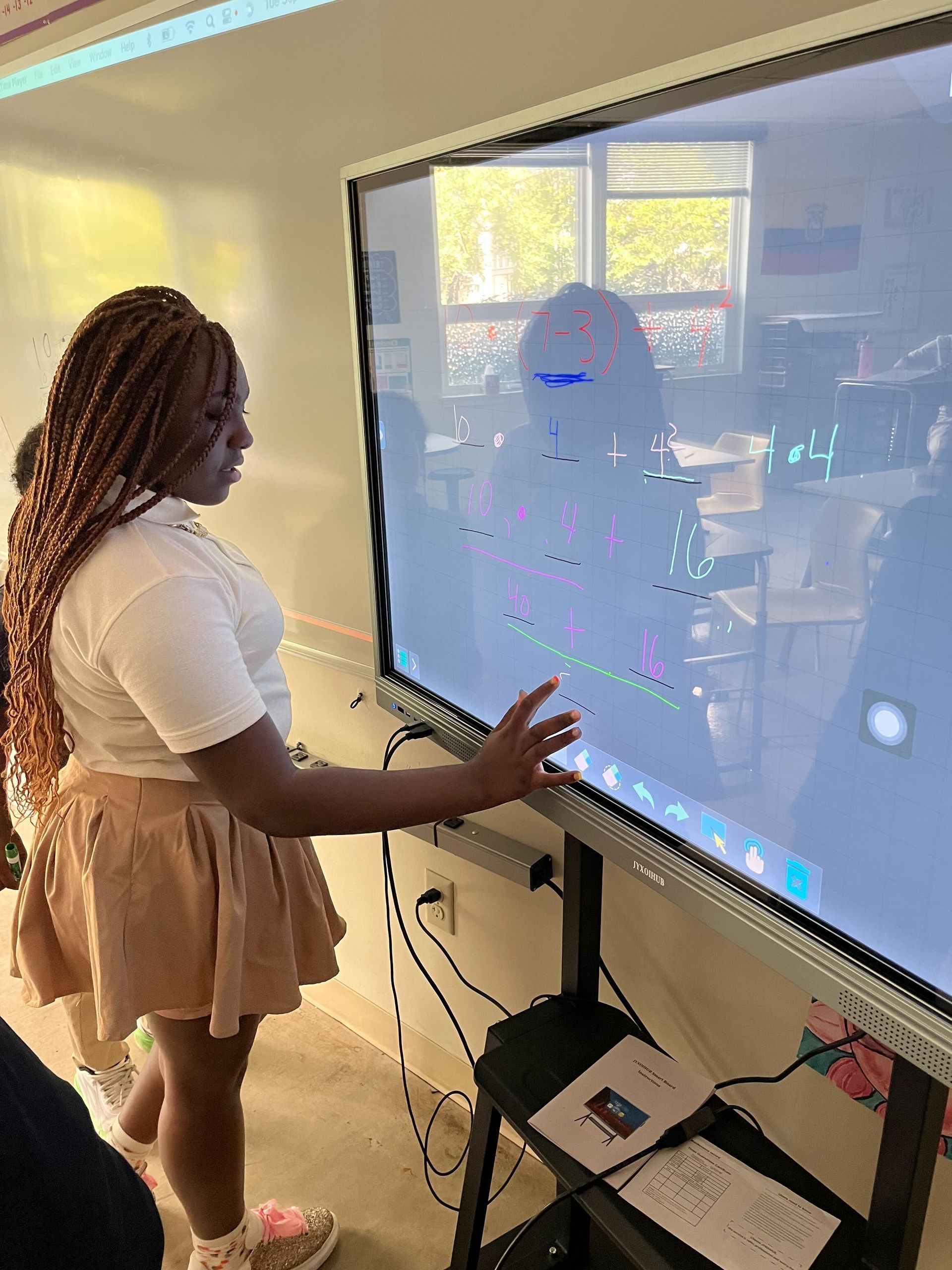 Crossroads School student points at a screen in a classroom, reflected in the display. Beige skirt, white shirt.