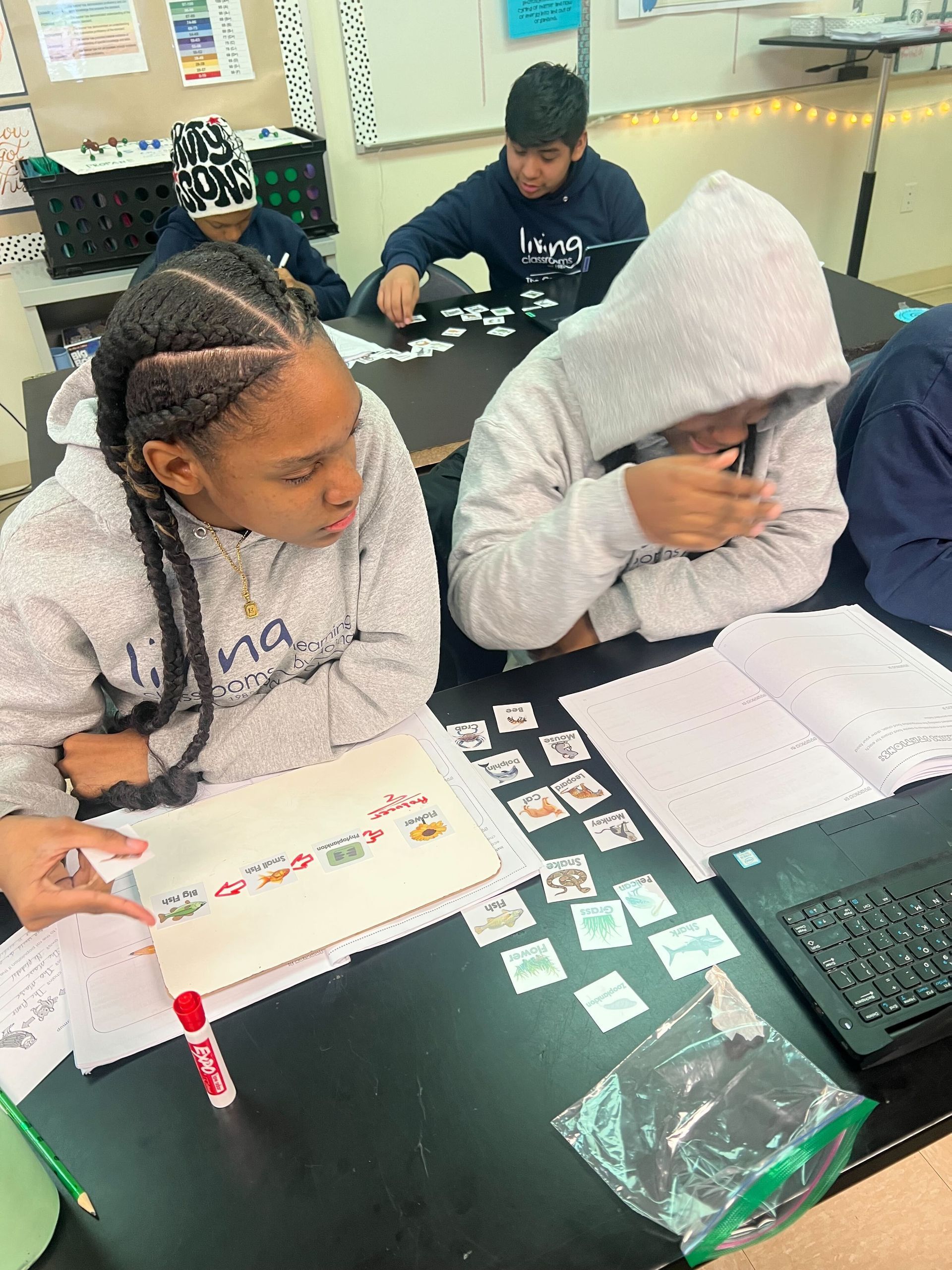 Crossroads School Students at a desk studying papers and cards. One student in a hoodie, a second student with braided hair. Notebooks and cards.