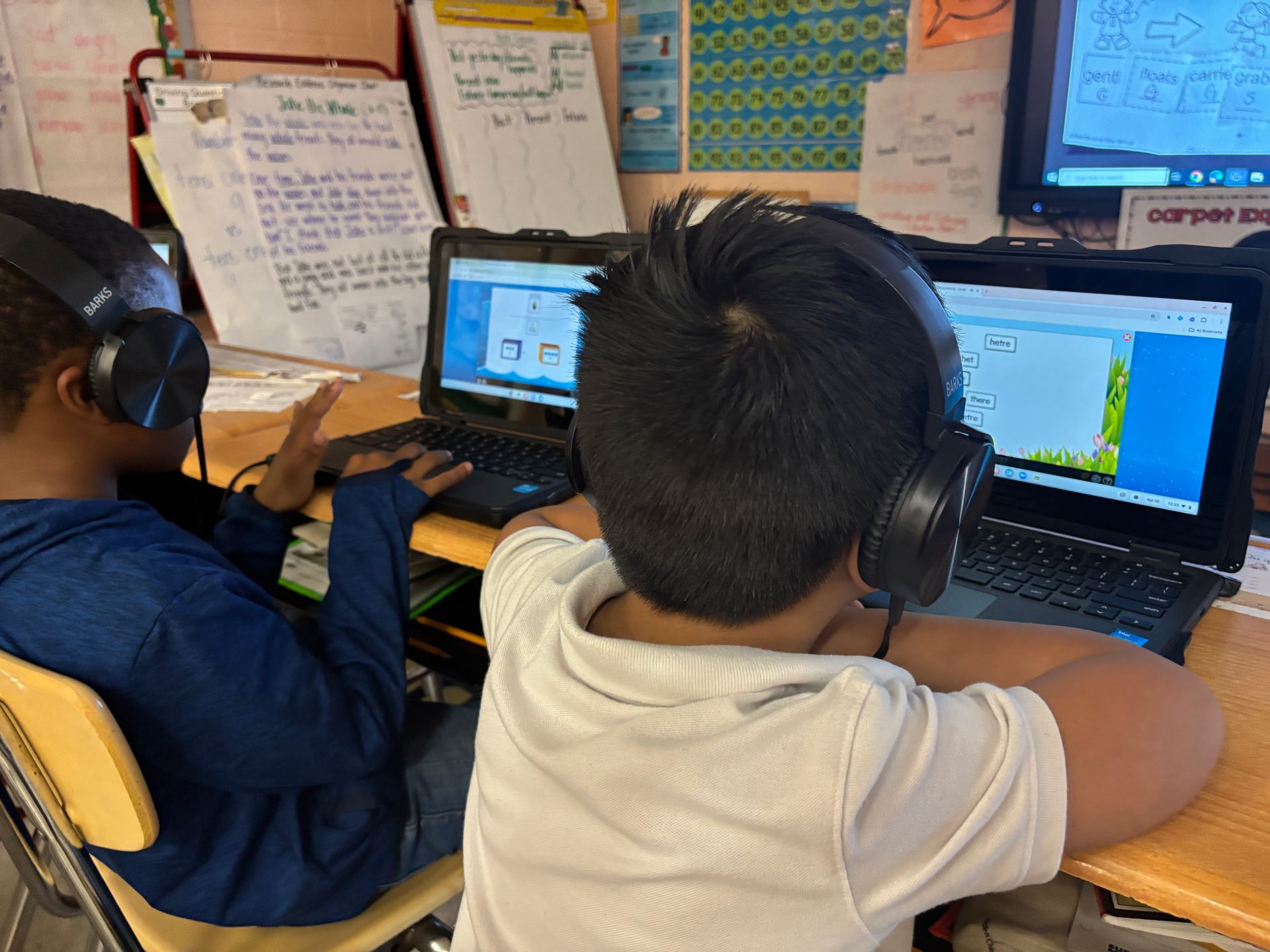 Two students wearing headphones, working on laptops at a desk in a classroom.