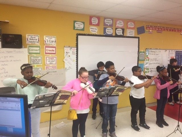 Children playing violins in a classroom, sheet music on stands, yellow wall, whiteboard.