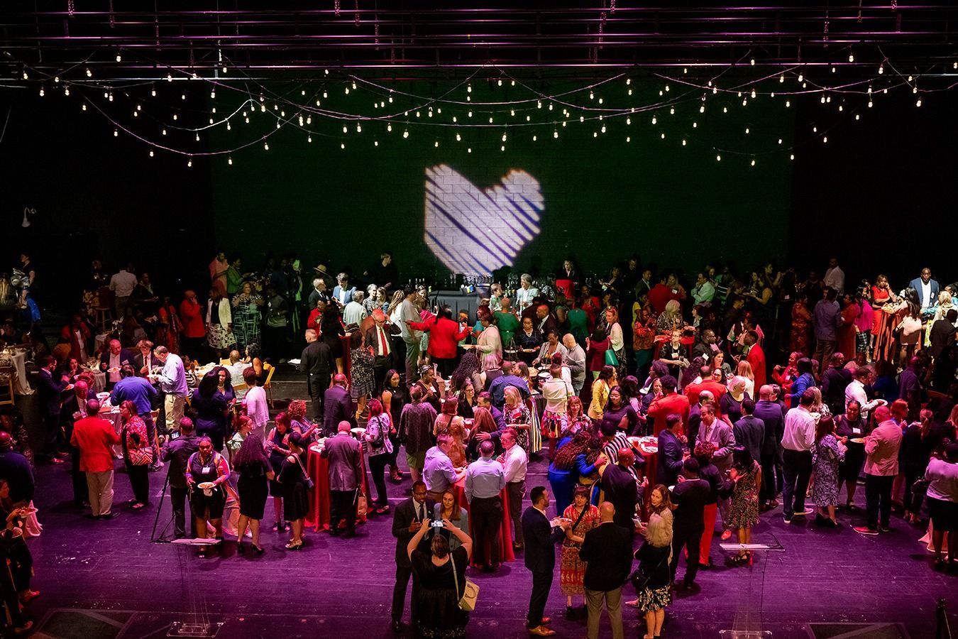 HOS Awards Ceremony - Large crowd at event with heart symbol on a green backdrop, under string lights.