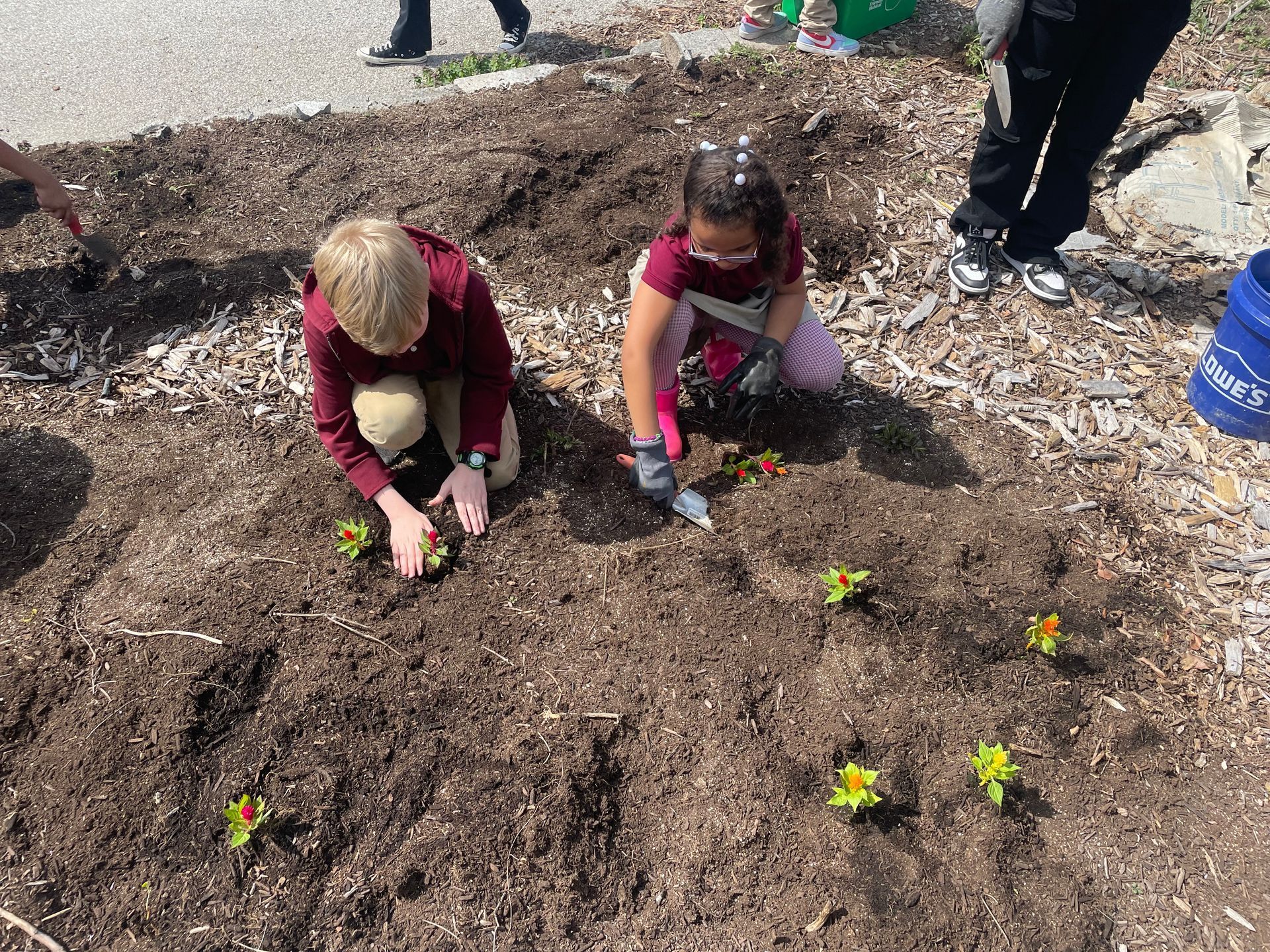 Children planting flowers in a garden bed, using trowels, outdoors.