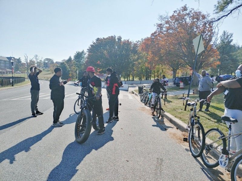 JCB Academy Bike Club - principals on bikes gathered on a paved road next to a grassy area with trees, under a blue sky.