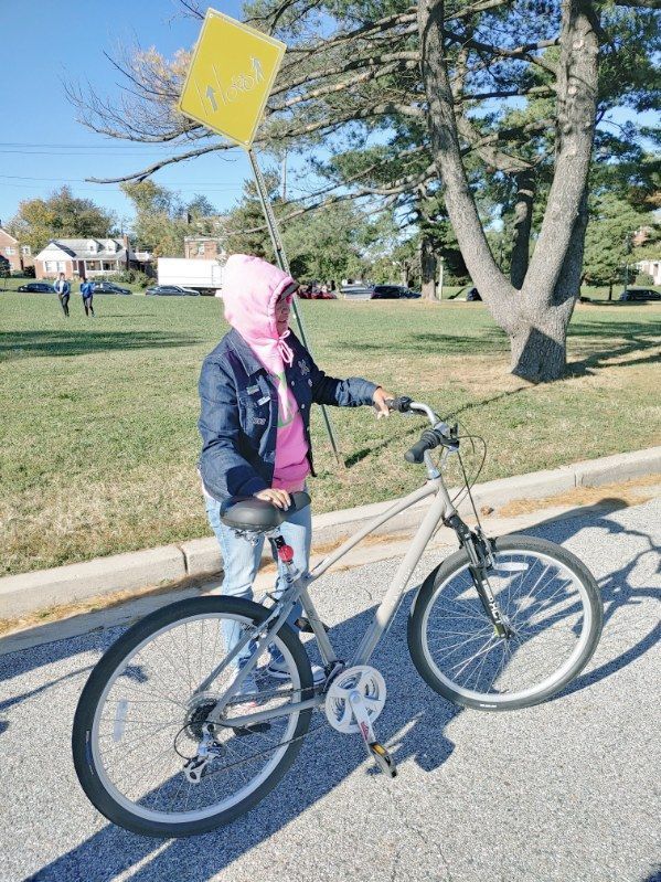 DC principal standing next to a bike, holding a yellow kite. Sunny day, outdoors.