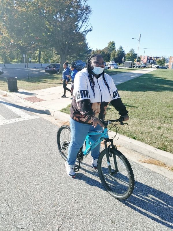 JCB Academy Bike Club - Principal riding a teal bike on a paved path. Wearing a mask, a black and white top, and jeans. Sunlight and green grass in the background.