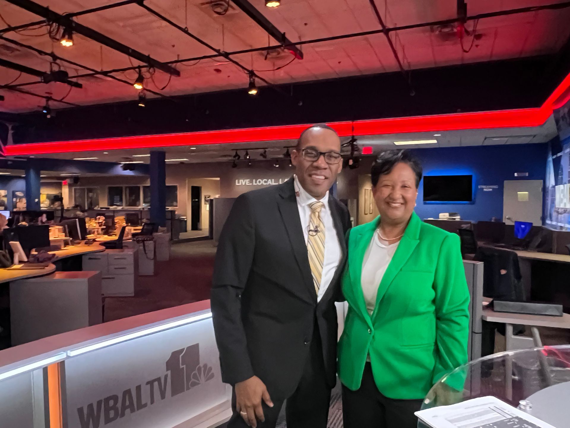 Two people smiling in a TV news studio. Man in suit, woman in green blazer, standing by a desk.