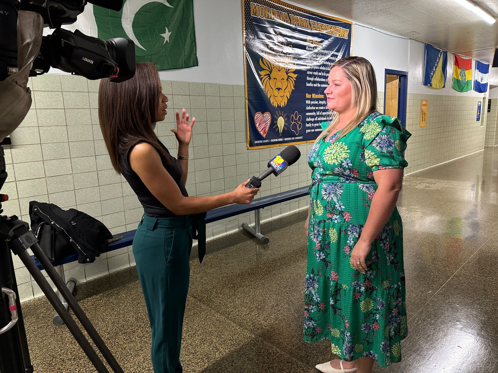 A reporter interviews a woman in a school hallway, holding a microphone. Pakistan flag is in the background.