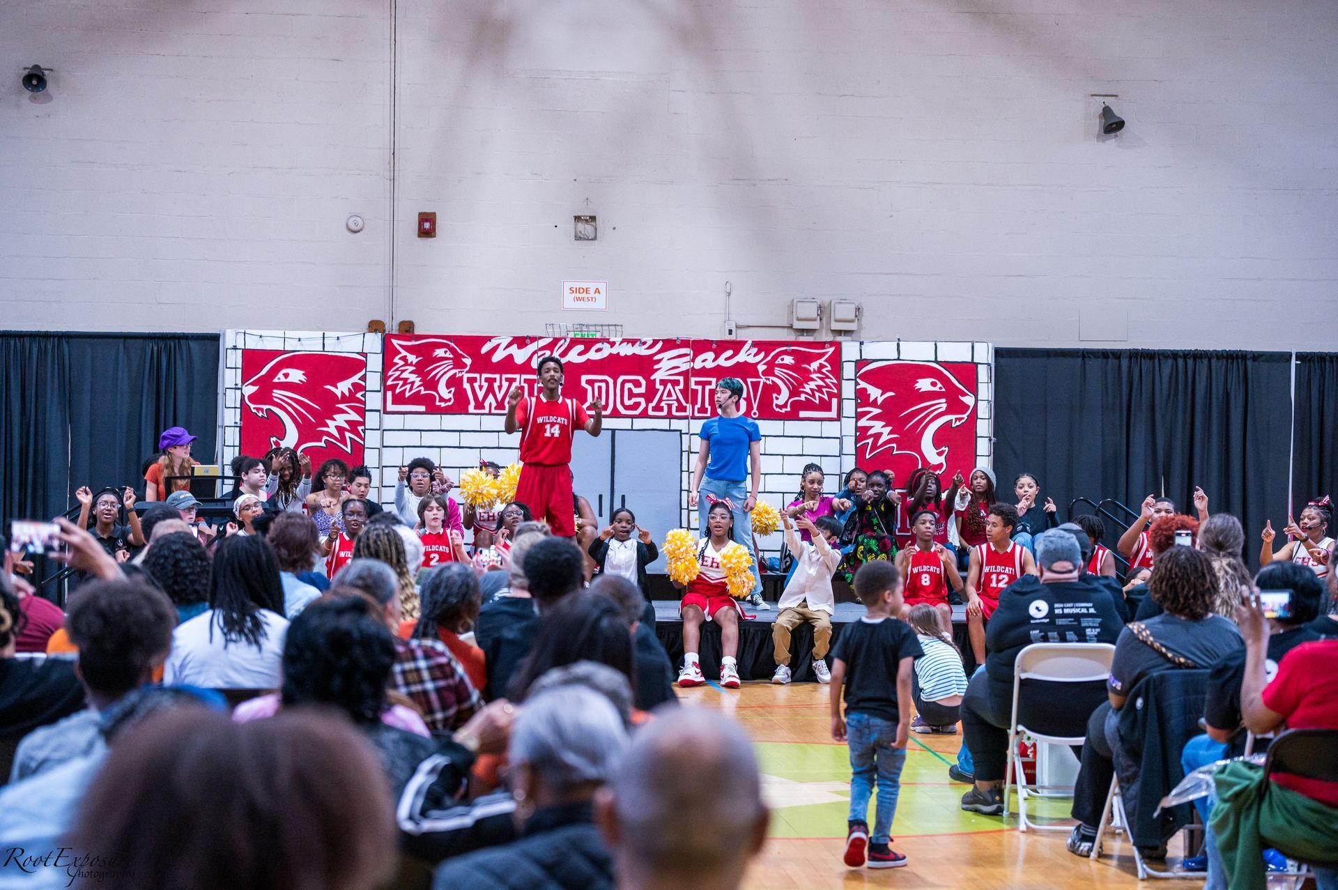Turnbridge School Pep Rally - Crowd watches performers on a stage in a gymnasium, banner in the background.