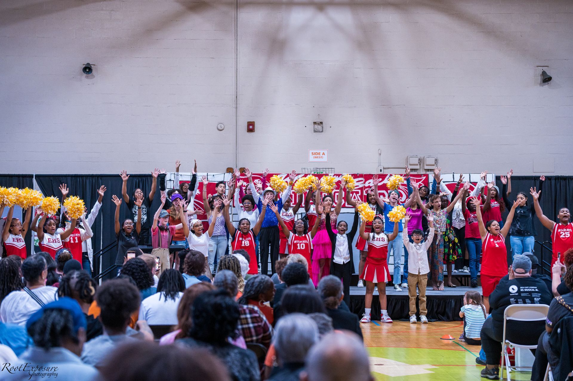 Turnbridge School - Group of people on stage with arms raised in a gymnasium. Some hold pom-poms. Audience seated in front.