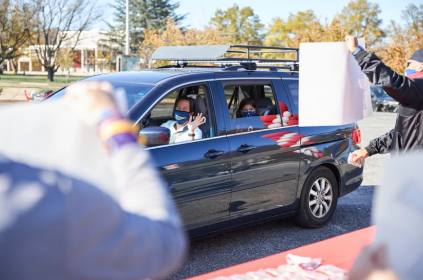 Drive Thru for You  Event - People in a car waving from the window, being handed signs by people outdoors on a sunny day.