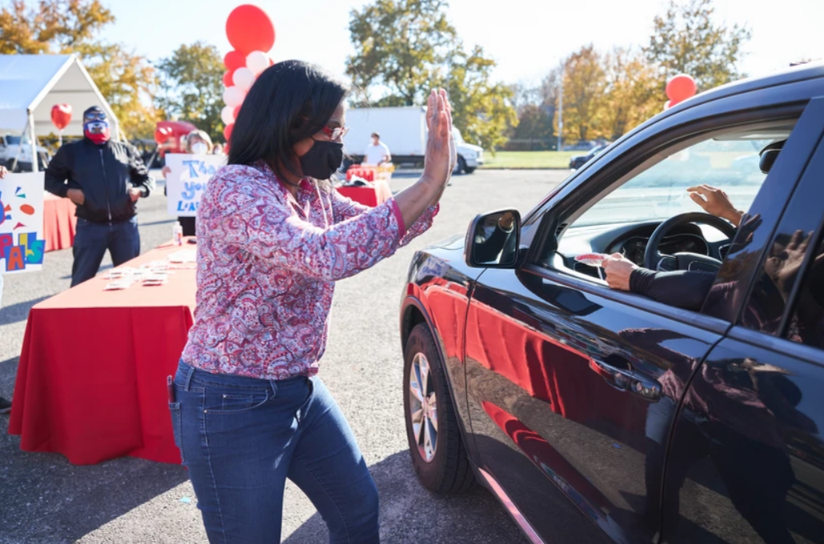 Woman in mask waves to person in car at outdoor event; red and white decor.