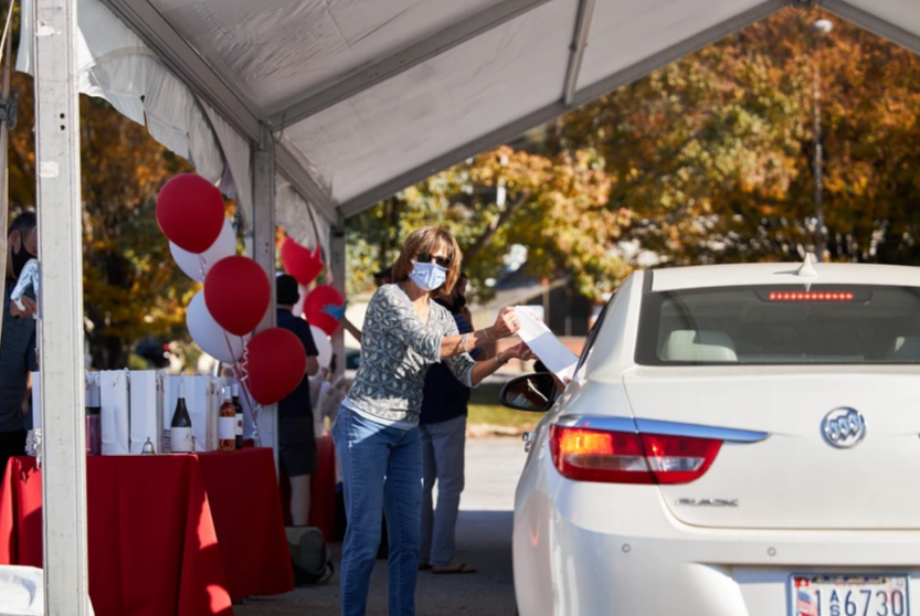 Drive Thru for You Event - Woman in mask hands document to driver in white car at outdoor event tent.