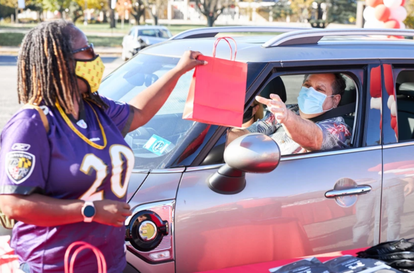 Drive Thru for You Event - Woman hands a red gift bag to a person in a car at a drive-through, both wearing masks.