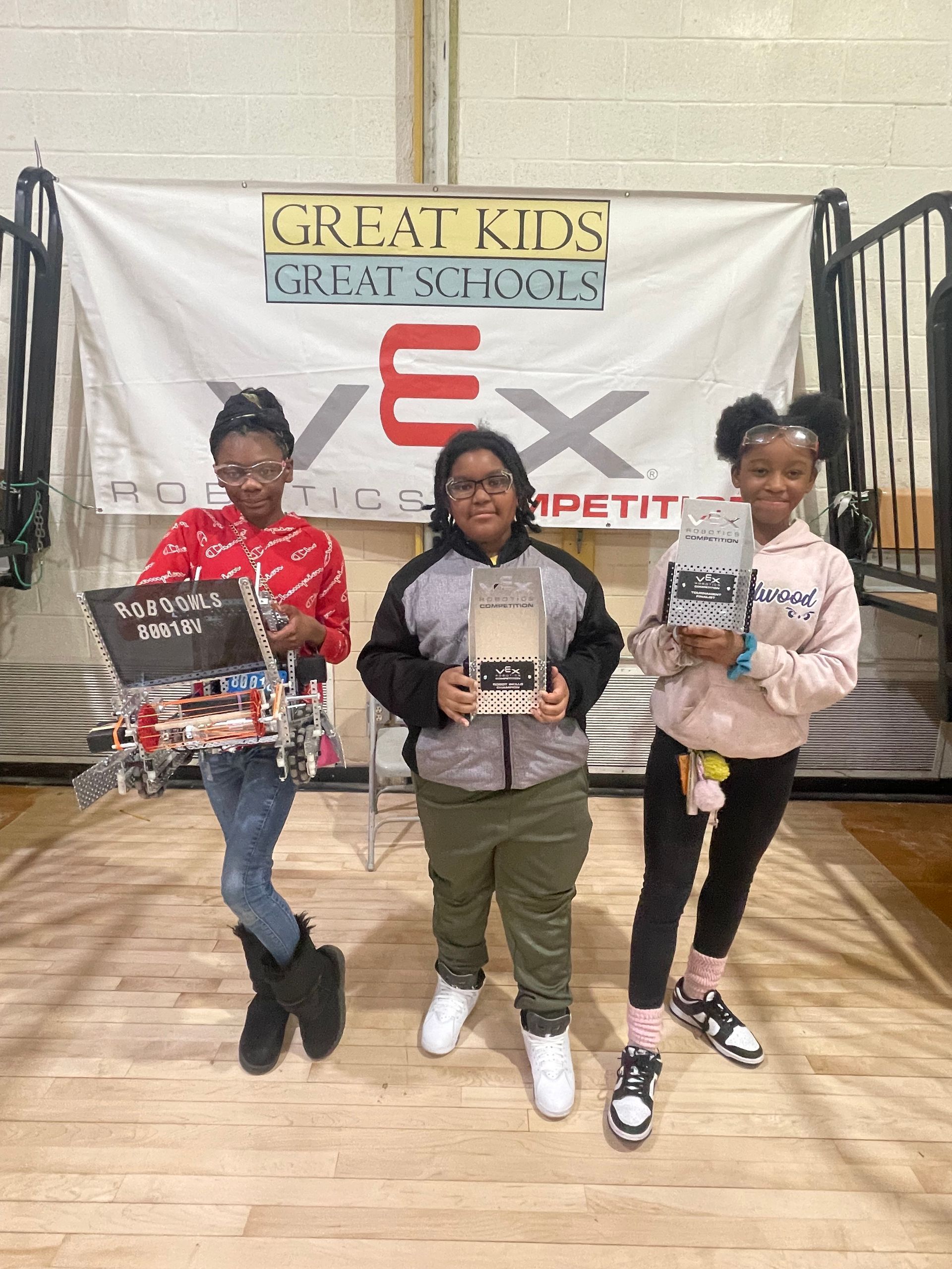 Hazelwood Elementary - Three students holding robots, posing in front of a VEX competition banner.