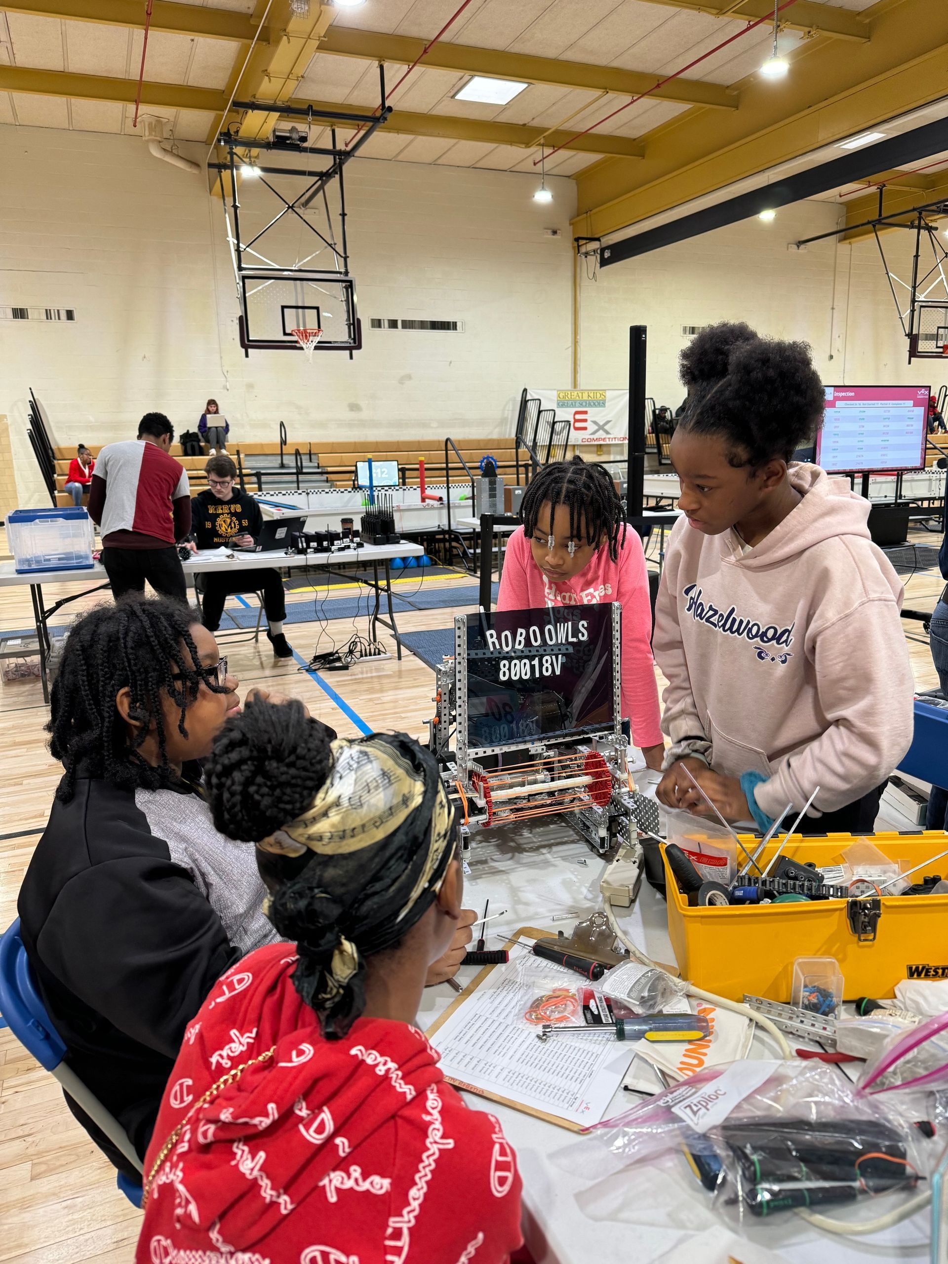 Hazelwood Elementary students working on a robot in a gymnasium. One girl in a pink sweater; others looking on. Tools and wires visible.
