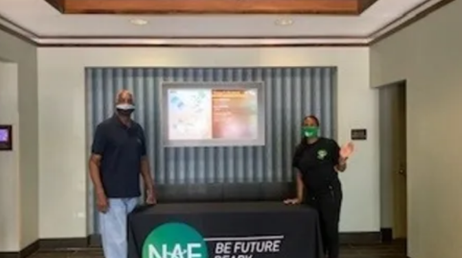 Path Forward Event - Two Principals stand behind a black table with a logo, in a lobby setting. One person waves.