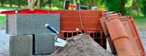 Materiales de construcción: bloques de hormigón, ladrillos, tejas, llana, nivel y pila de arena. Exterior con follaje verde.