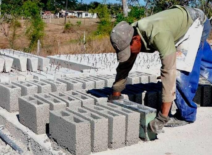 Trabajador apilando bloques de hormigón al aire libre. Camisa verde, pantalón azul y bloques grises.