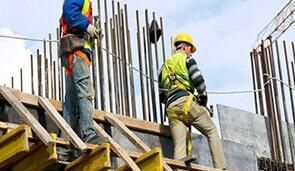 Obreros en una obra con arneses y cascos de seguridad. Hormigón y acero visibles.