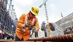 Obrero de la construcción con casco amarillo y chaleco naranja trabajando en una estructura metálica en un sitio de construcción.