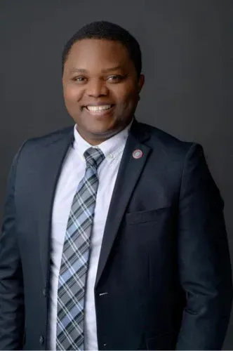 Man in a suit smiling, looking directly at the camera against a dark gray background.